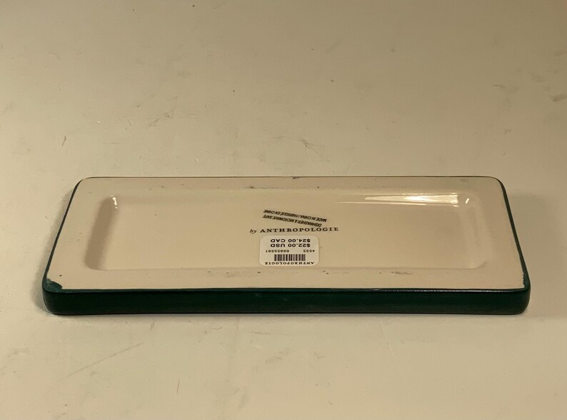 May include: Rectangular ceramic tray with a cream-colored interior and a dark green border. The tray has a slightly raised edge and a label that reads "by Anthropologie".
