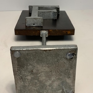 May include: A metal letter "F" sculpture. The letter is mounted on a dark brown wooden base. The base is supported by a metal rod attached to a square metal base. The sculpture has a rustic, industrial aesthetic.