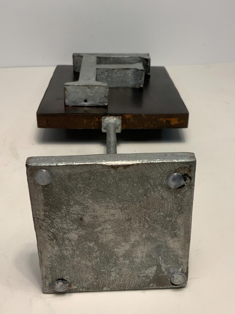 May include: A metal letter "F" sculpture. The letter is mounted on a dark brown wooden base. The base is supported by a metal rod attached to a square metal base. The sculpture has a rustic, industrial aesthetic.