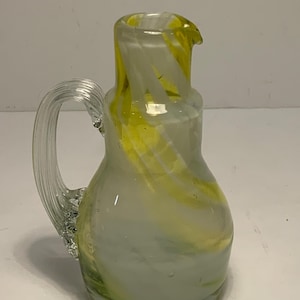 Może przedstawiać: Przezroczysty szklany dzbanek z uchwytem i wylewką. Dzbanek ma zaokrąglony korpus i rozszerzoną górę. Zawiera żółte i białe wiry w całym szkle. Uchwyt jest przezroczysty i zakrzywiony.