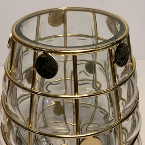 Op de afbeelding: Een heldere glazen vaas met een gouden metalen kooi-ontwerp. De vaas heeft een afgeronde vorm met een reeks cirkelvormige inkepingen. Goudkleurige cirkelvormige accenten zijn bevestigd aan de metalen kooi. De vaas is waarschijnlijk voor decoratieve doeleinden.