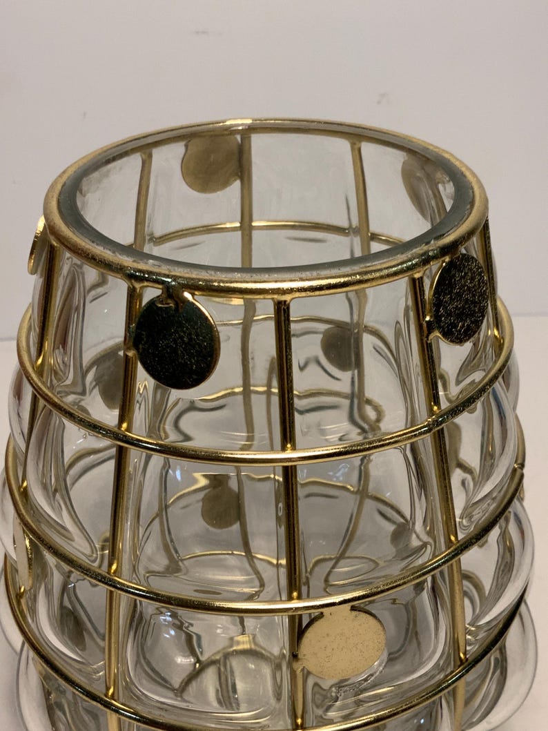 Op de afbeelding: Een heldere glazen vaas met een gouden metalen kooi-ontwerp. De vaas heeft een afgeronde vorm met een reeks cirkelvormige inkepingen. Goudkleurige cirkelvormige accenten zijn bevestigd aan de metalen kooi. De vaas is waarschijnlijk voor decoratieve doeleinden.