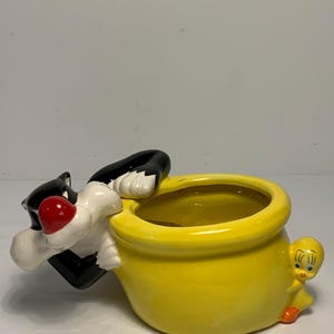 May include: A ceramic planter featuring Sylvester the Cat and Tweety Bird. Sylvester is black and white with a red nose, clinging to the edge of a bright yellow pot. Tweety Bird is yellow and orange, perched on the side of the pot.