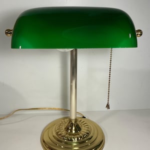 May include: A banker's lamp featuring a polished gold-tone metal base and a vertical fluted column. The lamp has a wide, curved emerald green glass shade with gold-tone side adjustment knobs and a gold-tone pull chain switch hanging from the right.