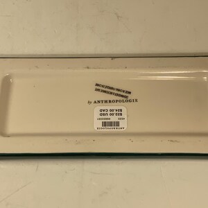 May include: A rectangular off-white Anthropologie dish with a green border. The dish has a sticker with the brand name, price, and other details. The dish is dishwasher and microwave safe.