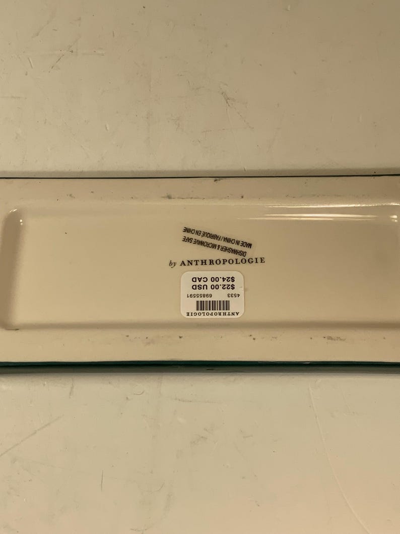 May include: A rectangular off-white Anthropologie dish with a green border. The dish has a sticker with the brand name, price, and other details. The dish is dishwasher and microwave safe.
