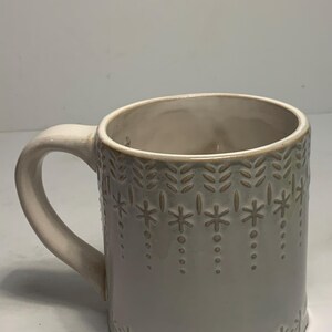 Peut inclure: Mug en c&eacute;ramique gris clair avec une anse cr&egrave;me. Le mug pr&eacute;sente un motif en relief de flocons de neige et de motifs de feuilles. L'int&eacute;rieur est beige clair. Un design simple et &eacute;l&eacute;gant pour un usage quotidien. Id&eacute;al pour les boissons chaudes ou froides.