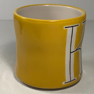 Puede incluir: Una taza de cer&aacute;mica amarilla con interior blanco y una gran letra "K" blanca en el lateral. La taza tiene un acabado liso y brillante y una forma redondeada. La letra "K" est&aacute; delineada en negro.