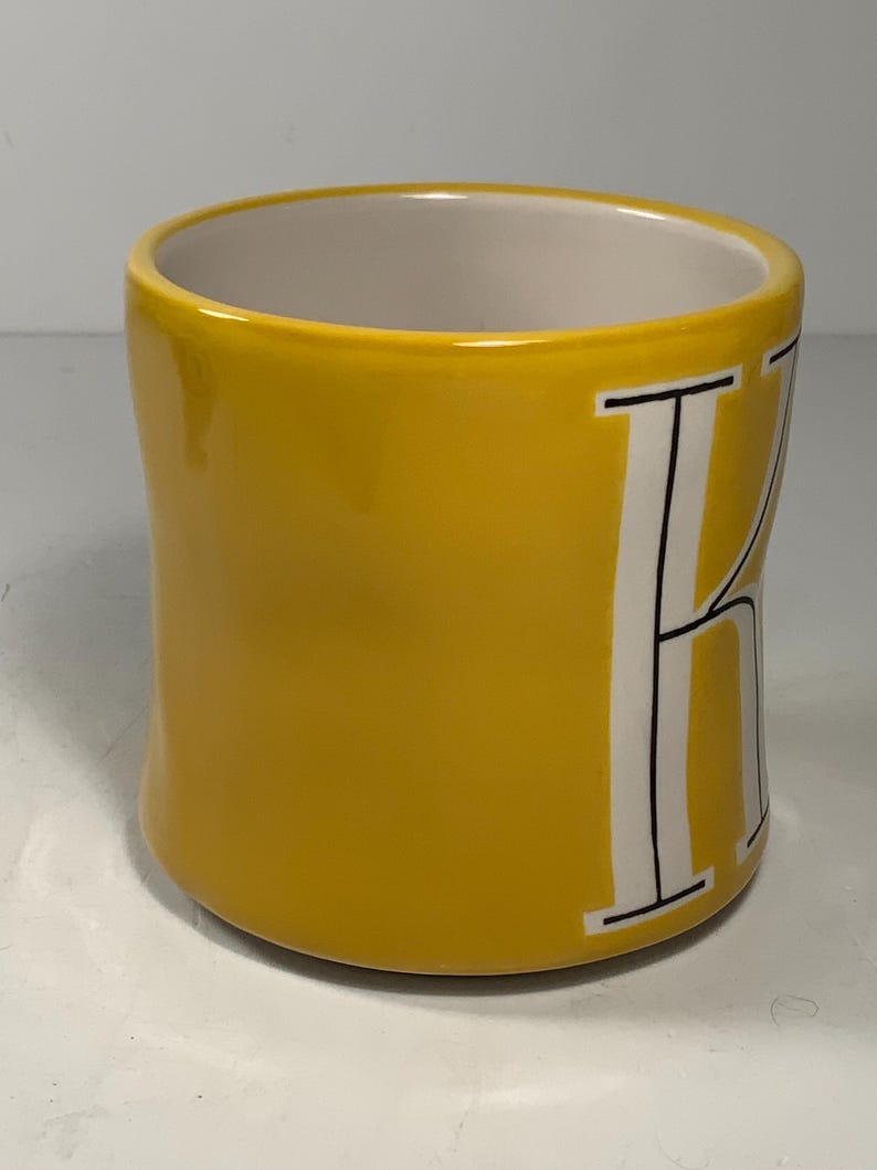 Puede incluir: Una taza de cer&aacute;mica amarilla con interior blanco y una gran letra "K" blanca en el lateral. La taza tiene un acabado liso y brillante y una forma redondeada. La letra "K" est&aacute; delineada en negro.