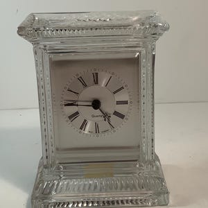 Puede incluir: Un reloj de cuarzo de cristal transparente con forma rectangular. La esfera es blanca con números romanos y manecillas negras. Las palabras "Quartz" y "West Germany" están impresas en la esfera. El reloj tiene un diseño decorativo y texturizado.