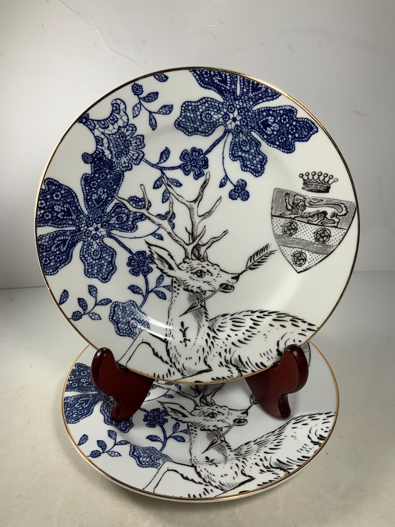 May include: Two white ceramic plates with a blue and gold floral design and a black and white deer illustration. The plates have a gold rim.