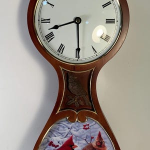 May include: Wooden wall clock with a round face featuring Roman numerals and black hands. The clock has a decorative lower section with a winter scene of red cardinals. The clock is approximately 20 inches tall.