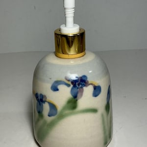 May include: A ceramic soap dispenser with a rounded body featuring a hand-painted floral pattern of blue irises with green stems on an off-white background. The dispenser is topped with a gold-colored collar and a white plastic pump mechanism.