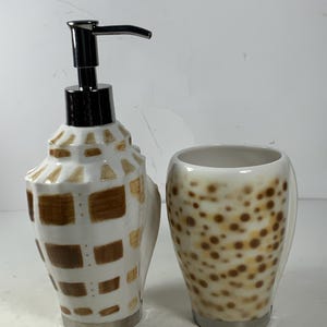 Puede incluir: Un juego de dispensador de jabón y vaso de cerámica blanca con un diseño de concha marrón. El dispensador tiene una bomba cromada y el vaso tiene un diseño moteado marrón y blanco.