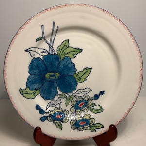 May include: A white ceramic plate with a blue floral design. The plate features a large blue flower with green leaves and smaller blue and yellow flowers. The rim has a scalloped red border. The plate is displayed on a wooden stand.
