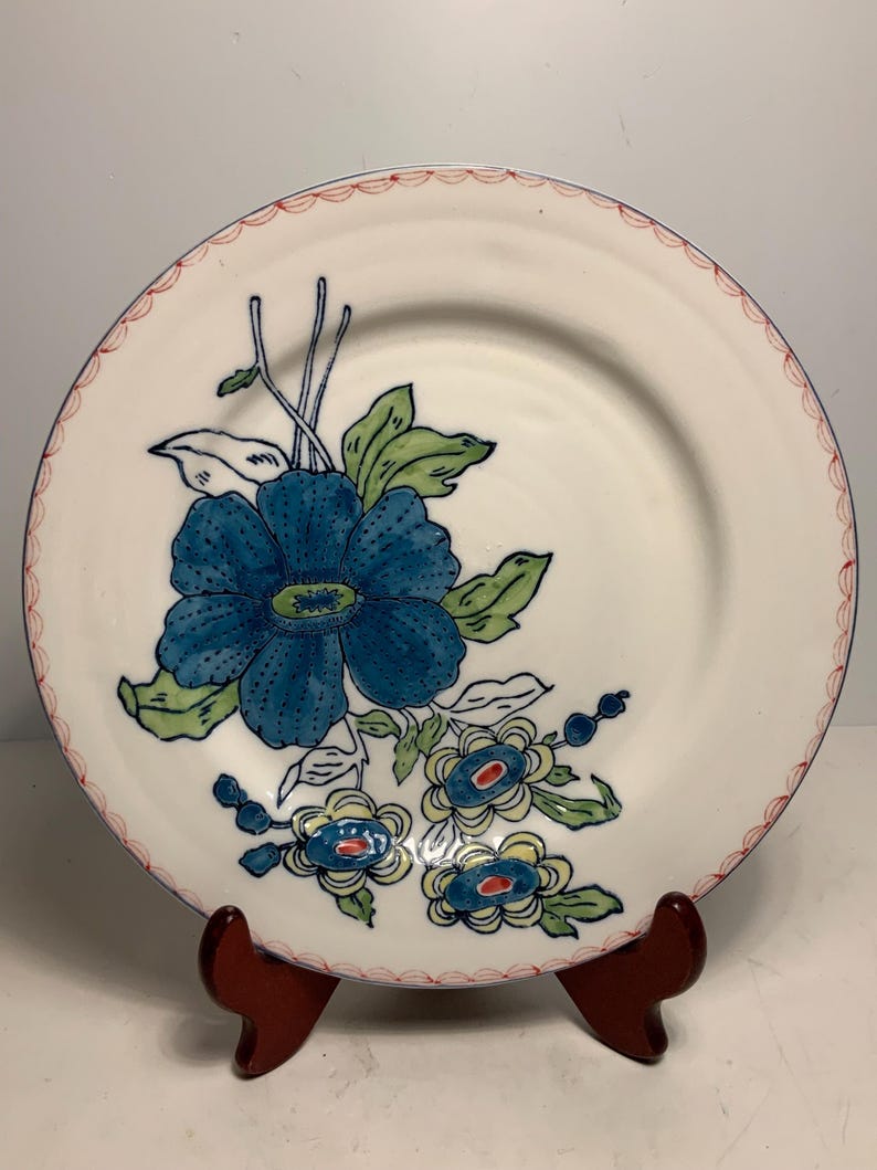 May include: A white ceramic plate with a blue floral design. The plate features a large blue flower with green leaves and smaller blue and yellow flowers. The rim has a scalloped red border. The plate is displayed on a wooden stand.