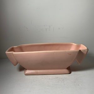 Pode incluir: Um vaso de cerâmica rosa claro com formato retangular e duas alças de cada lado. O vaso tem um acabamento liso e brilhante.