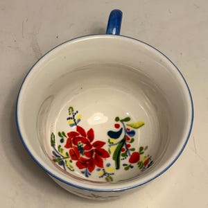 May include: White ceramic bowl with a blue handle and rim, decorated with a floral pattern in red, yellow, and green. The bowl's interior features a vibrant floral design, adding a touch of color to the white background. A charming piece for serving or display.