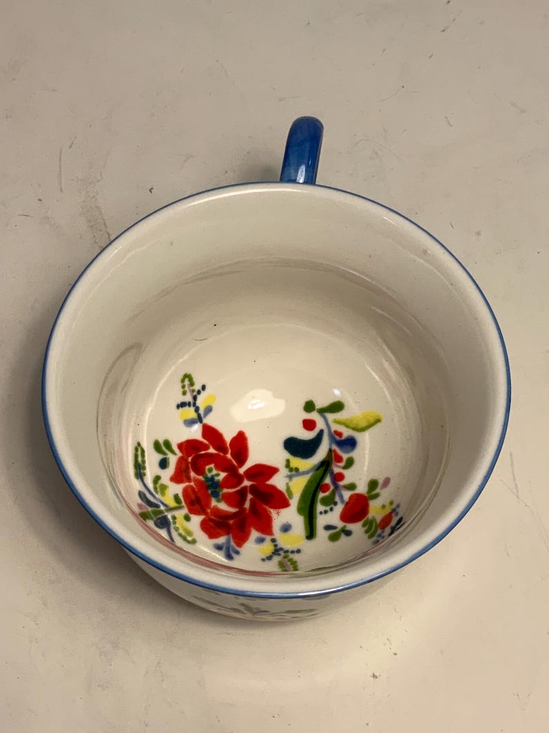 May include: White ceramic bowl with a blue handle and rim, decorated with a floral pattern in red, yellow, and green. The bowl's interior features a vibrant floral design, adding a touch of color to the white background. A charming piece for serving or display.