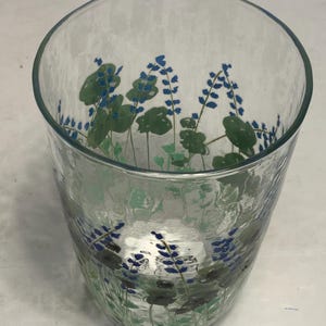 May include: A clear glass tumbler with a textured surface. The glass is decorated with a floral design featuring blue flowers and green leaves. The tumbler has a wide opening and a slightly curved shape.