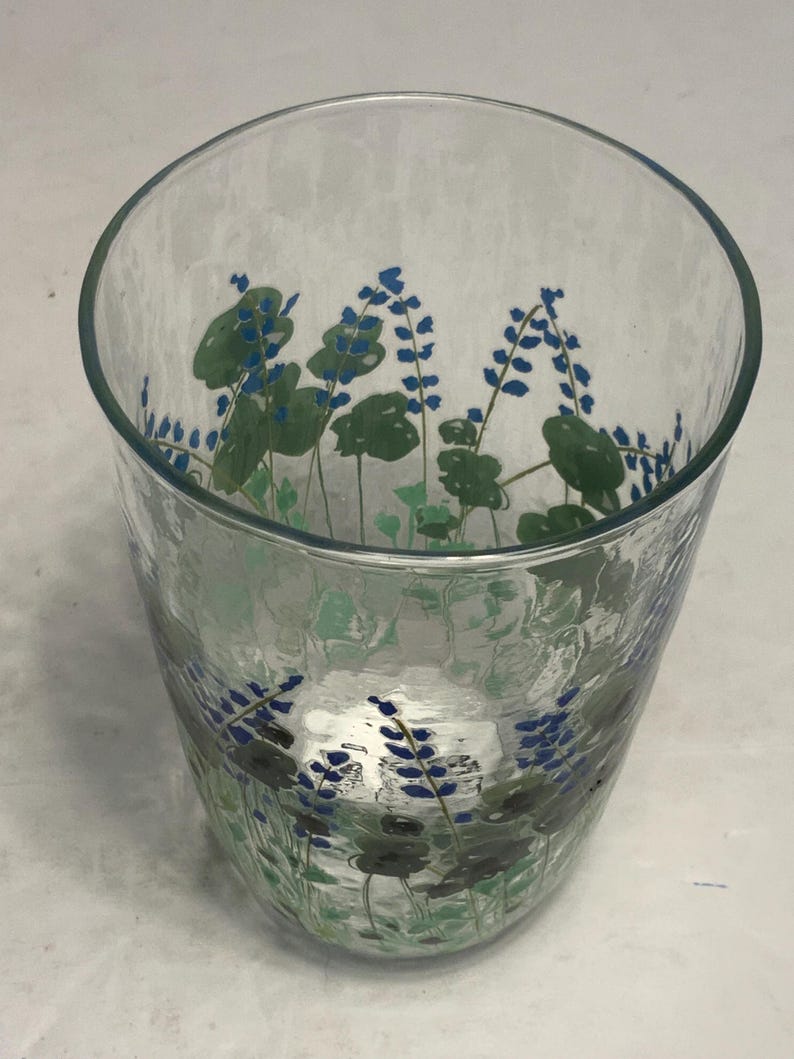 May include: A clear glass tumbler with a textured surface. The glass is decorated with a floral design featuring blue flowers and green leaves. The tumbler has a wide opening and a slightly curved shape.