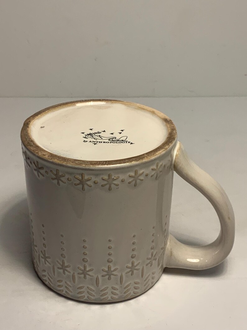 Peut inclure: Une tasse en c&eacute;ramique cr&egrave;me avec un motif d&eacute;coratif de flocons de neige et de points. La tasse a une anse incurv&eacute;e et le texte "ANTHROPOLOGIE" est visible en bas.