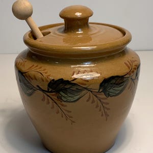 May include: A tan ceramic honey pot with a matching lid and a wooden honey dipper. The pot is decorated with a leaf and vine design in shades of green and brown. The pot is round with a small handle on the lid.