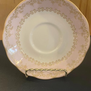 May include: A vintage teacup saucer with a scalloped edge. The saucer has a pale pink border with gold filigree and a gold rim. The centre is white. The saucer is displayed on a gold-coloured metal stand.