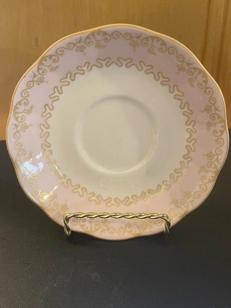 May include: A vintage teacup saucer with a scalloped edge. The saucer features a pale pink border with gold filigree and a gold rim. The center is white. The saucer is displayed on a gold-colored metal stand.