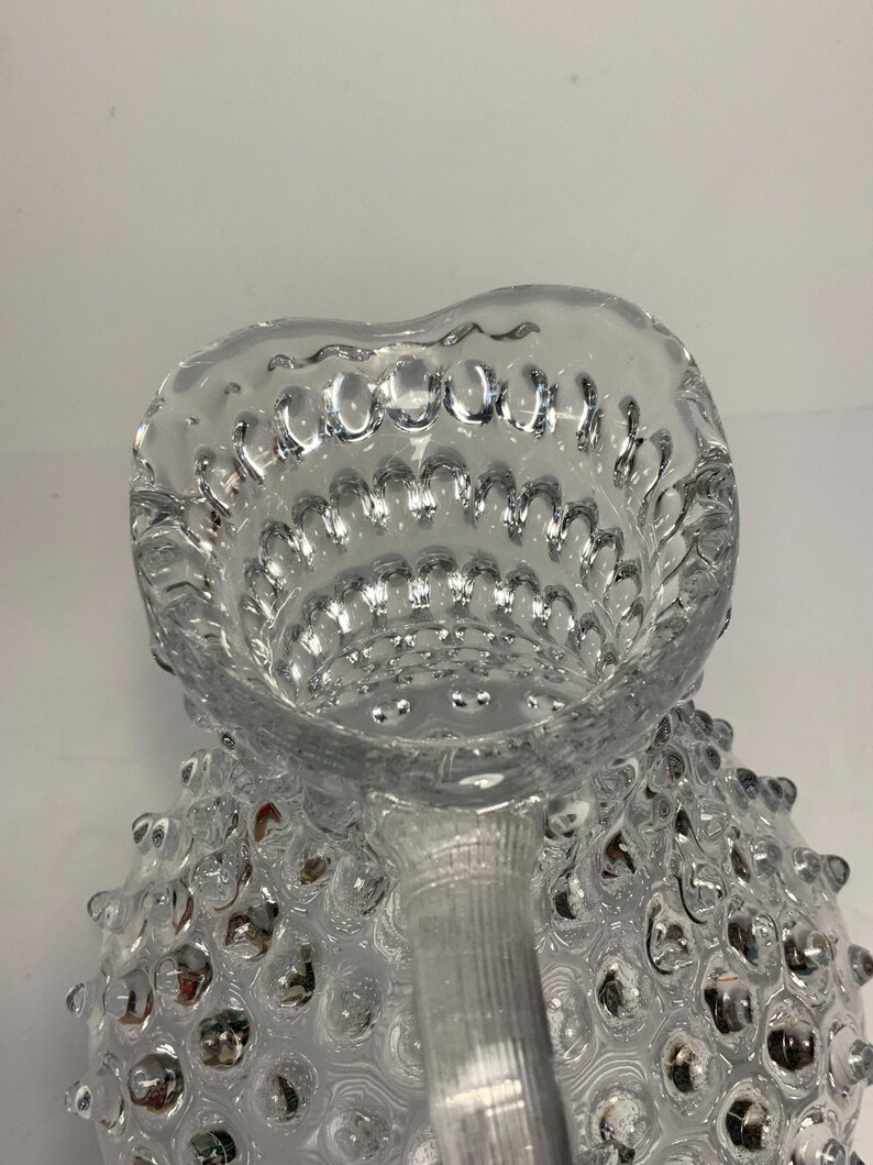May include: Clear glass pitcher with a textured, bubble-like pattern. The pitcher has a heart-shaped opening and a handle. The glass is transparent, allowing light to pass through, and the design adds visual interest.