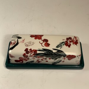 May include: A rectangular butter dish with a cream-colored lid featuring a floral design in red and black. The base is a dark green. The dish is designed to hold butter.