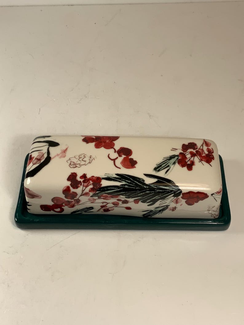 May include: A rectangular butter dish with a cream-colored lid featuring a floral design in red and black. The base is a dark green. The dish is designed to hold butter.