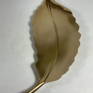 Puede incluir: Una decoraci&oacute;n de pared en forma de hoja dorada con una superficie texturizada. La hoja es curva y tiene un tallo largo.