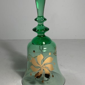 May include: A green glass bell with a long handle and a flared base. The bell is decorated with a gold floral design. The clapper hangs inside the bell. The bell is made of transparent glass.