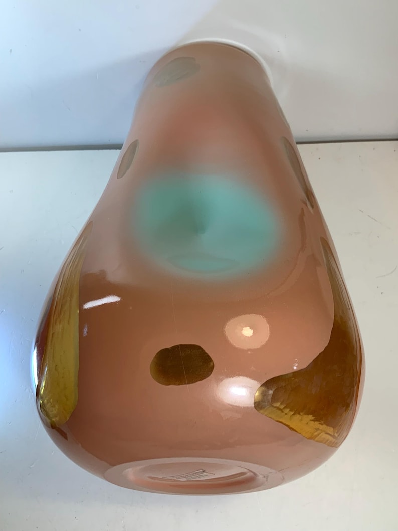 May include: A tall, teardrop-shaped vase with a peach-colored body. The vase features irregular brown and gold accents, with a central turquoise-colored area. The glass vase has a glossy finish and a wide base.