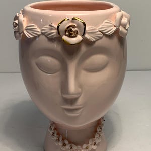 May include: A pink ceramic face vase with closed eyes, a floral crown, and a floral necklace. The crown features white roses with gold accents. The vase is a decorative item for home decor.