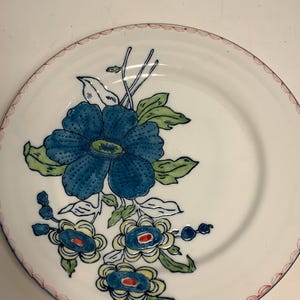May include: White ceramic plate with a blue floral design. The plate features a large blue flower with green leaves and smaller blue and yellow flowers. A red scalloped border trims the edge.