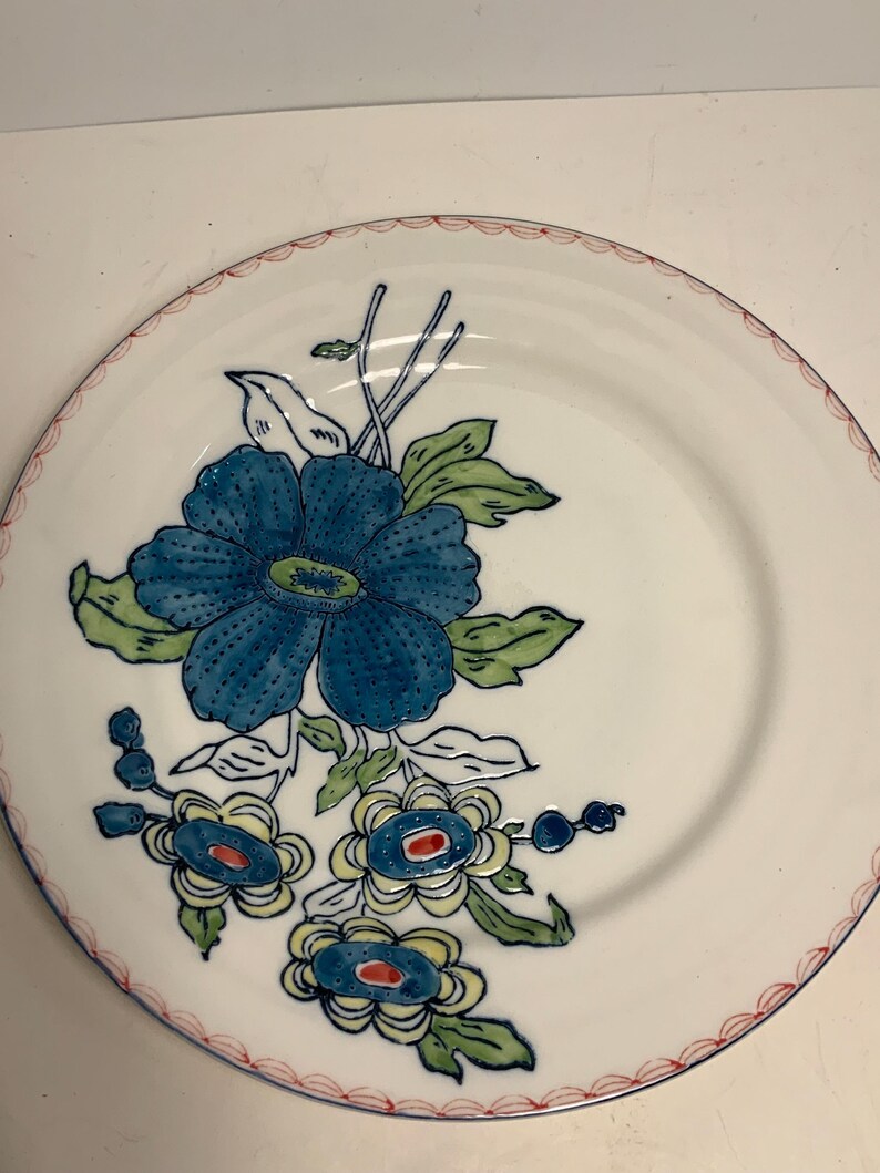 May include: White ceramic plate with a blue floral design. The plate features a large blue flower with green leaves and smaller blue and yellow flowers. A red scalloped border trims the edge.