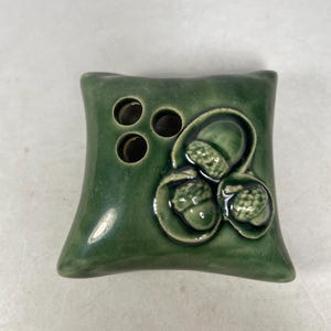 May include: A green ceramic pillow-shaped vase with three holes for flowers and an acorn design.
