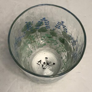 May include: Clear glass tumbler with a textured surface and a blue rim. The glass is decorated with blue and green floral designs. The bottom of the glass has the letters "IA" and a black floral design.