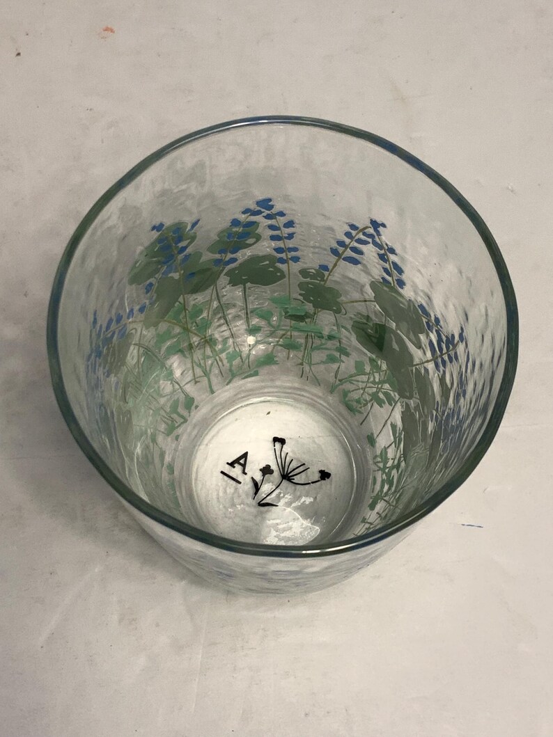 May include: Clear glass tumbler with a textured surface and a blue rim. The glass is decorated with blue and green floral designs. The bottom of the glass has the letters "IA" and a black floral design.