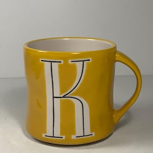 Puede incluir: Una taza de cer&aacute;mica amarilla con interior blanco y asa curva. La letra "K" se muestra en blanco con contorno negro en la parte delantera. La taza est&aacute; sobre un fondo blanco liso.