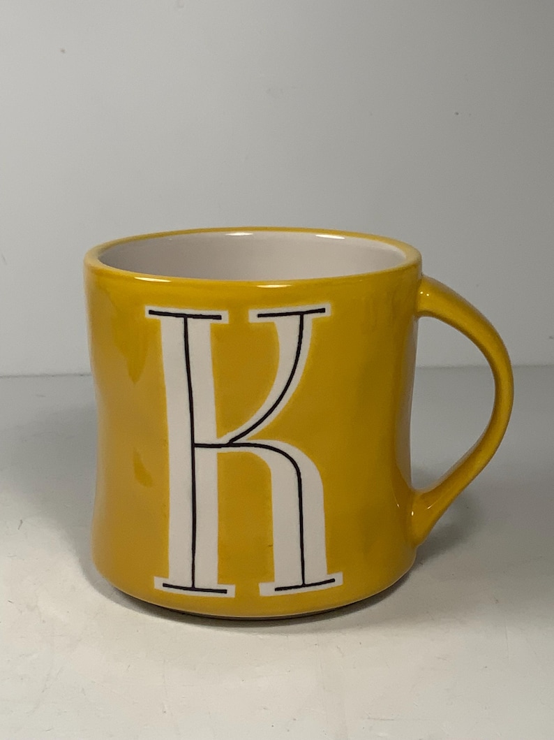 Puede incluir: Una taza de cer&aacute;mica amarilla con interior blanco y asa curva. La letra "K" se muestra en blanco con contorno negro en la parte delantera. La taza est&aacute; sobre un fondo blanco liso.
