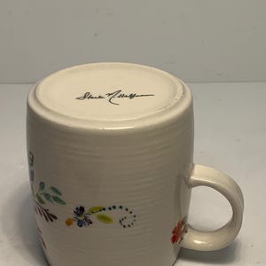 Puede incluir: Taza de cer&aacute;mica color crema con un dise&ntilde;o floral en rojo, azul y verde. La taza tiene un asa curva y la firma del artista en la parte inferior. La taza mide aproximadamente 10 cm de alto.
