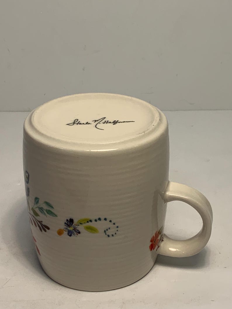 Puede incluir: Taza de cer&aacute;mica color crema con un dise&ntilde;o floral en rojo, azul y verde. La taza tiene un asa curva y la firma del artista en la parte inferior. La taza mide aproximadamente 10 cm de alto.