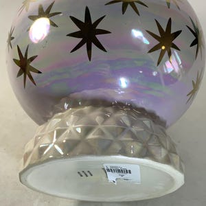 May include: A pearlescent, spherical decorative vase with gold star accents. The vase has a textured, geometric base and a wide, white opening. The iridescent finish reflects a range of colors, creating a celestial aesthetic.