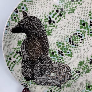 May include: A decorative ceramic plate featuring a black and white fox illustration. The plate has a white background with a speckled pattern and green and black accents. The fox is detailed with intricate patterns.