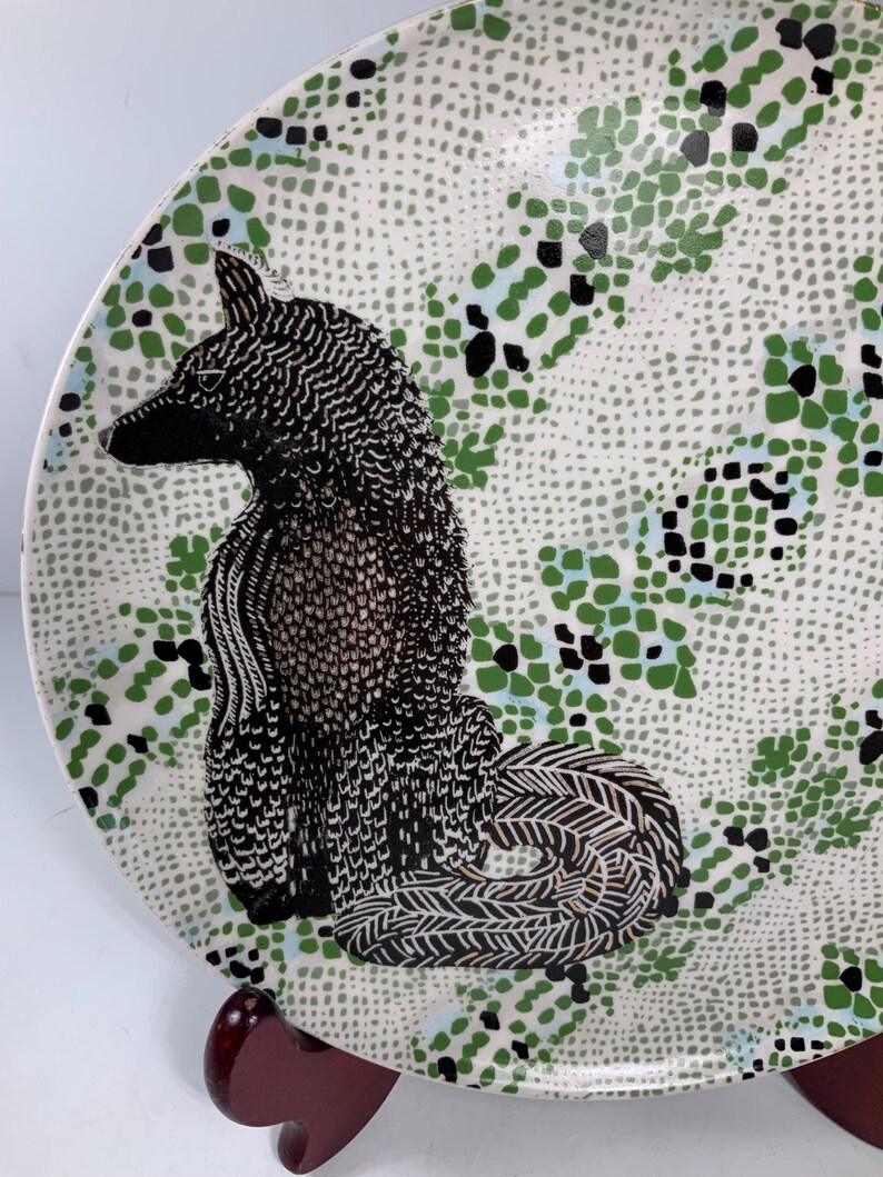 May include: A decorative ceramic plate featuring a black and white fox illustration. The plate has a white background with a speckled pattern and green and black accents. The fox is detailed with intricate patterns.