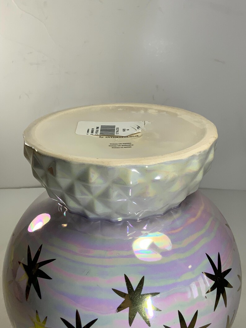 May include: Iridescent decorative vase with a round base and a textured, geometric top. The vase features gold starburst designs. The bottom of the vase is a pearlescent purple, and the top is a light gray.