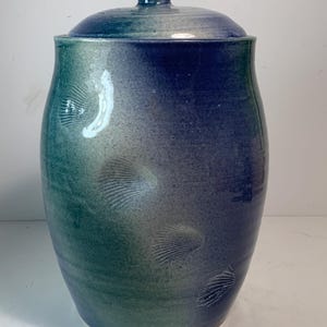 May include: A handcrafted ceramic jar with a lid, featuring a gradient of blue and green hues. The jar has a cylindrical shape with embossed shell-like patterns. The lid has a small knob for easy handling. A decorative and functional piece.